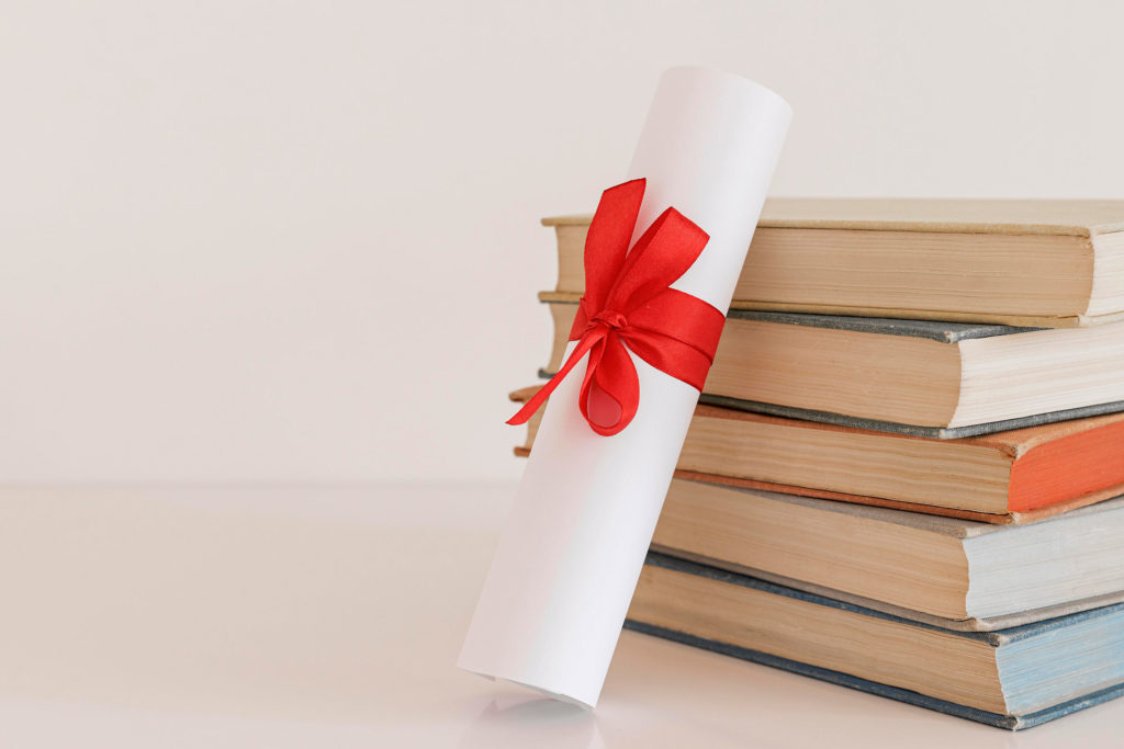 "Stack of books with a rolled diploma tied with a red ribbon on top, placed on a white background."