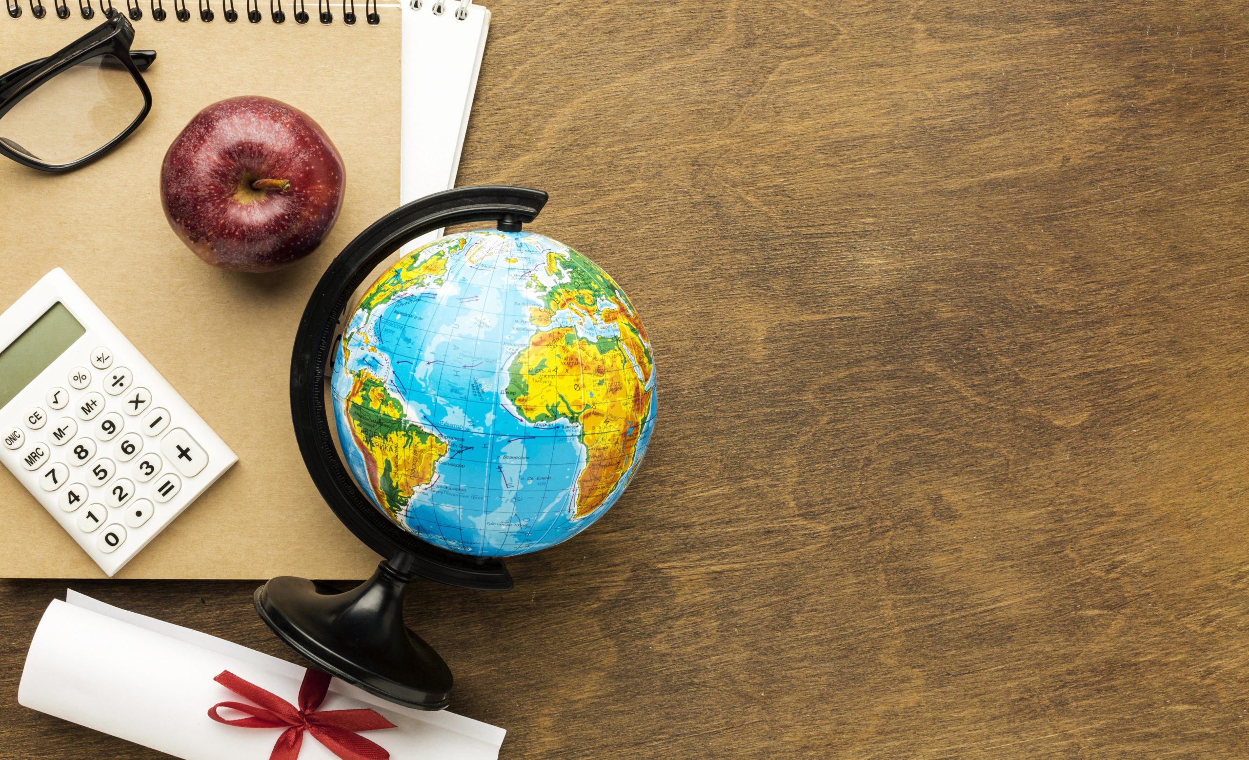 "Items on a wooden desk including a globe, calculator, red apple, glasses, and a notebook."