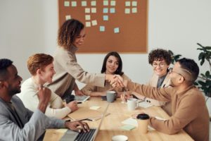 A diverse group of professionals sits around a table in a modern office, as a woman and a man shake hands with smiles.