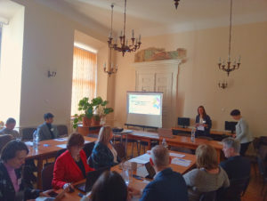 People sitting at tables in a meeting room with a presentation being given. A projector screen displays a slide.