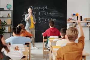 A teacher in a gray blazer and yellow shirt gestures while standing in front of a blackboard with math equations, as students sit at desks, one raising their hand.