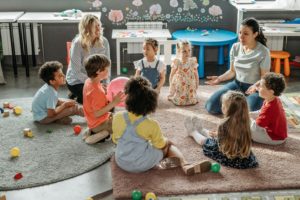 Children sit in a circle on the floor with two adults in a classroom setting, surrounded by toys and colorful tables.