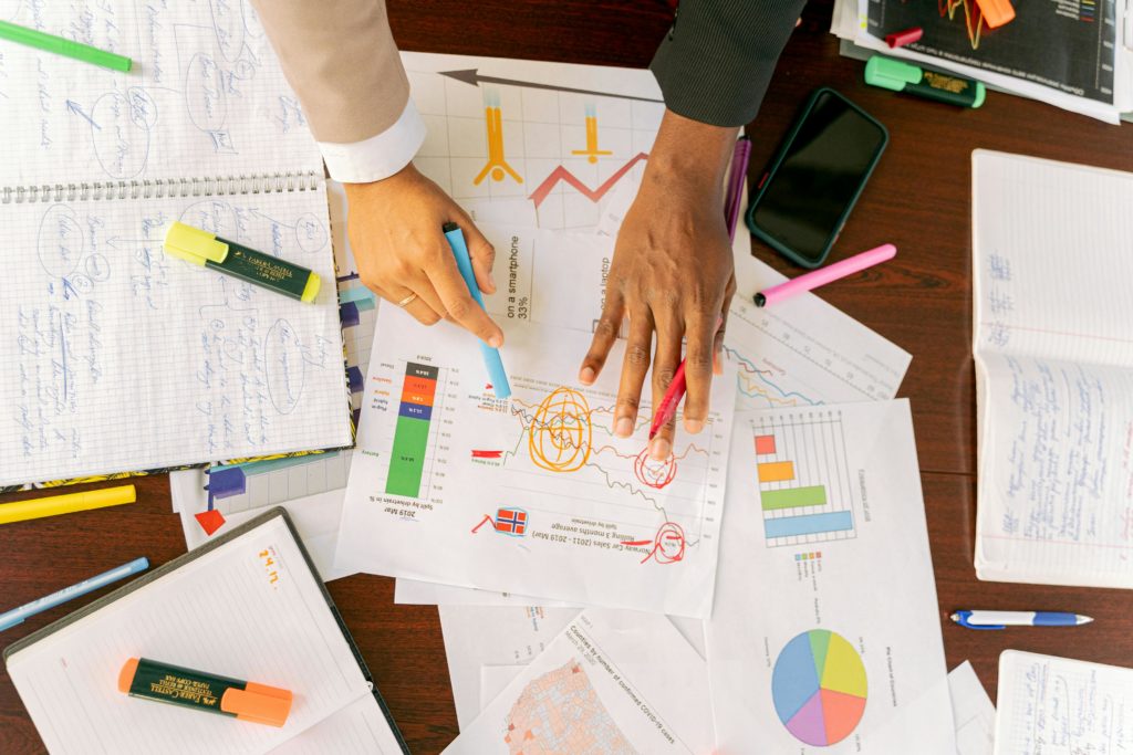 Two people pointing at graphs and charts on a cluttered table with notebooks, markers, and papers.