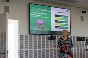 picture of the speaker during the conference: A woman stands in a well-lit room beside a large screen displaying a slide titled “Girls’ Empowerment in Politics – 5 Class Intervention,” which includes an illustrated graphic of diverse students and a list of five political learning questions. The presenter is wearing a floral dress and appears to be mid-speech.