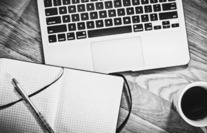 Black and white photo of a laptop, notebook, pen, and coffee cup on a wooden table.