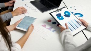 A group of people collaborating on a laptop and tablet, with graphs and charts displayed on the table in front of them.