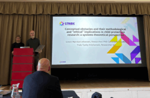 A man is giving a presentation at a conference, addressing an audience with visual aids in the background.