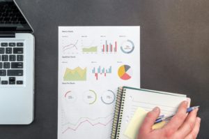 hand holding a pen over a notebook on a desk with a laptop and graphs in the background
