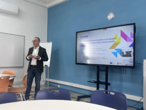 A man presents in a classroom, standing beside a large screen displaying a slide about teacher competence and student learning outcomes.