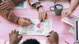 Team meeting with diverse professionals reviewing printed financial reports and colorful pie charts on a desk, using pens and a laptop, with a coffee cup nearby in a modern office setting.