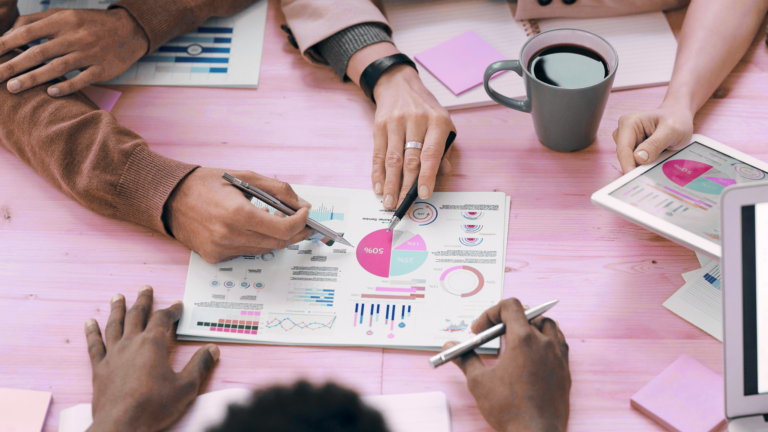 Team meeting with diverse professionals reviewing printed financial reports and colorful pie charts on a desk, using pens and a laptop, with a coffee cup nearby in a modern office setting.
