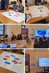 A workshop setting with people engaged in collaborative discussions around tables. Posters, laptops, and a presentation screen display colorful charts and text, indicating an academic or strategic planning context. The atmosphere is focused and interactive.